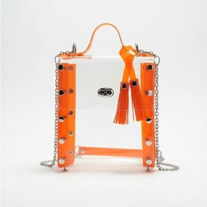 May include: A clear, rectangular handbag with bright orange accents. The bag features a top handle, chain strap, and decorative tassels. Orange panels are secured with silver rivets. The bag has a silver clasp and a clear body.
