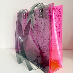 Puede incluir: Un bolso tote transparente con un dise&ntilde;o degradado rosa y morado. El bolso presenta asas de color gris oscuro y est&aacute; decorado con purpurina rosa y un patr&oacute;n moteado. El bolso est&aacute; hecho de un material transparente.