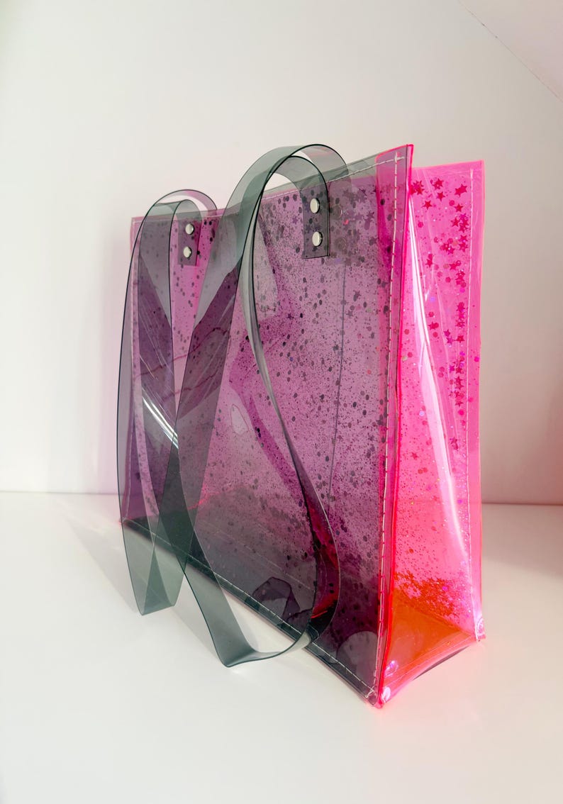 Puede incluir: Un bolso tote transparente con un dise&ntilde;o degradado rosa y morado. El bolso presenta asas de color gris oscuro y est&aacute; decorado con purpurina rosa y un patr&oacute;n moteado. El bolso est&aacute; hecho de un material transparente.