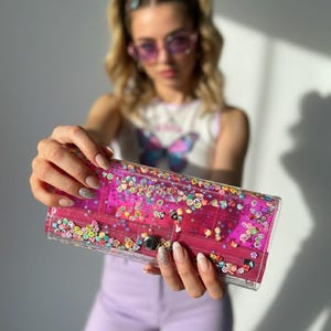 May include: A clear, rectangular clutch with a pink interior and a silver clasp. The clutch is filled with colorful, decorative shapes. The clutch is held up in front of a person.