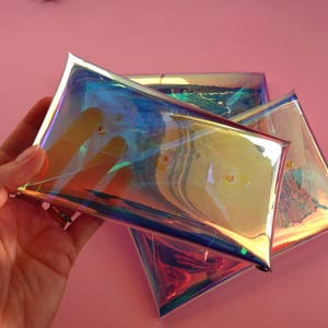 May include: Three iridescent clear plastic pouches stacked on top of each other. The pouches are made of a holographic material that reflects light in a rainbow of colors.