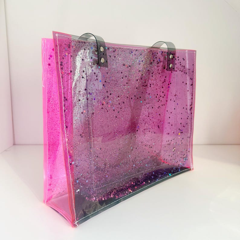 Puede incluir: Un bolso tote transparente con un dise&ntilde;o de purpurina rosa. El bolso tiene forma rectangular con un borde rosa y asas negras. El interior est&aacute; lleno de purpurina brillante.