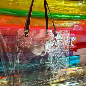 Puede incluir: Un bolso tote rectangular y transparente hecho de material pl&aacute;stico. El bolso cuenta con dos correas de cuero marr&oacute;n oscuro sujetas con remaches de metal plateado. Un bolsillo rectangular est&aacute; cosido en la parte exterior frontal.