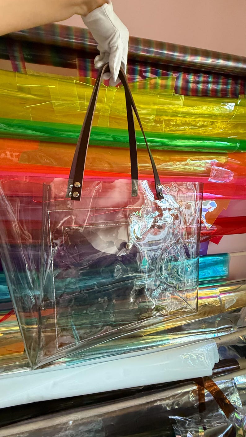 Puede incluir: Un bolso tote rectangular y transparente hecho de material pl&aacute;stico. El bolso cuenta con dos correas de cuero marr&oacute;n oscuro sujetas con remaches de metal plateado. Un bolsillo rectangular est&aacute; cosido en la parte exterior frontal.