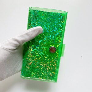 May include: A clear, rectangular, green glitter wallet with a silver snap closure. The wallet is filled with sparkling green glitter. The wallet is held by a gloved hand against a white background.