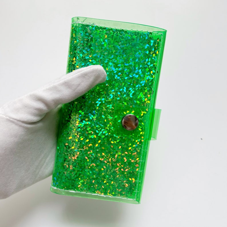 May include: A clear, rectangular, green glitter wallet with a silver snap closure. The wallet is filled with sparkling green glitter. The wallet is held by a gloved hand against a white background.