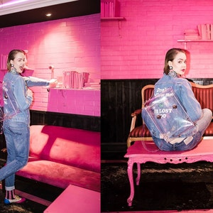 May include: A clear backpack with the text "I'M LOST 2020" and "Don't follow me" is worn with a denim jacket and jeans. The background is a pink room with pink furniture and a shelf with books. The overall theme is fashion.