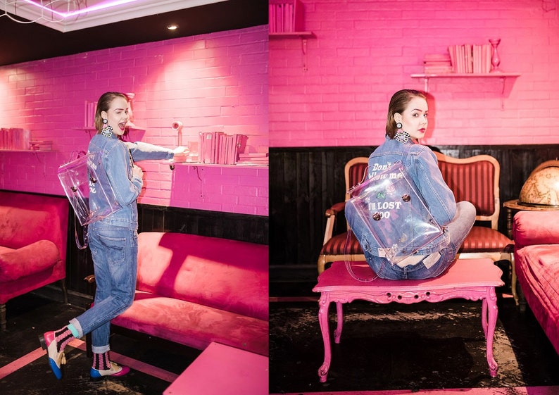 May include: A clear backpack with the text "I'M LOST 2020" and "Don't follow me" is worn with a denim jacket and jeans. The background is a pink room with pink furniture and a shelf with books. The overall theme is fashion.