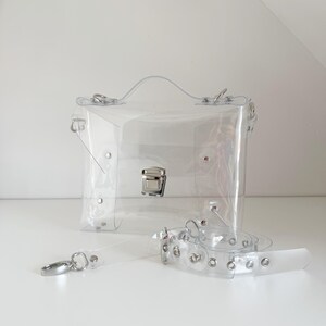 May include: A clear, rectangular handbag with a silver clasp and handle. The bag includes a detachable clear strap with silver hardware. The bag is made of transparent material, allowing visibility of the contents.