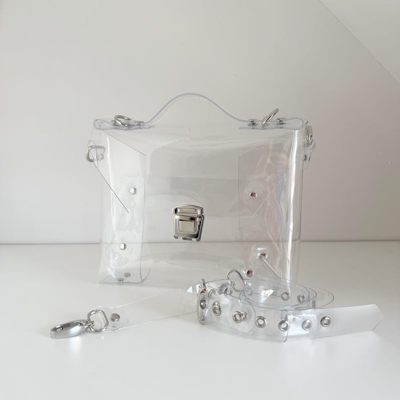 May include: A clear, rectangular handbag with a silver clasp and handle. The bag includes a detachable clear strap with silver hardware. The bag is made of transparent material, allowing visibility of the contents.
