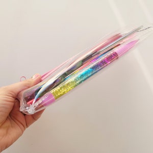 Pu&ograve; includere: Un astuccio trasparente e iridescente contenente varie penne e strumenti colorati. Le penne hanno fusti pieni di glitter nei toni del rosa, blu e giallo. Un cordino rosa &egrave; attaccato all'astuccio.