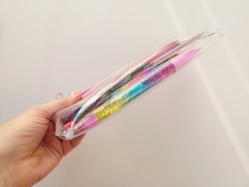 Pu&ograve; includere: Un astuccio trasparente e iridescente contenente varie penne e strumenti colorati. Le penne hanno fusti pieni di glitter nei toni del rosa, blu e giallo. Un cordino rosa &egrave; attaccato all'astuccio.