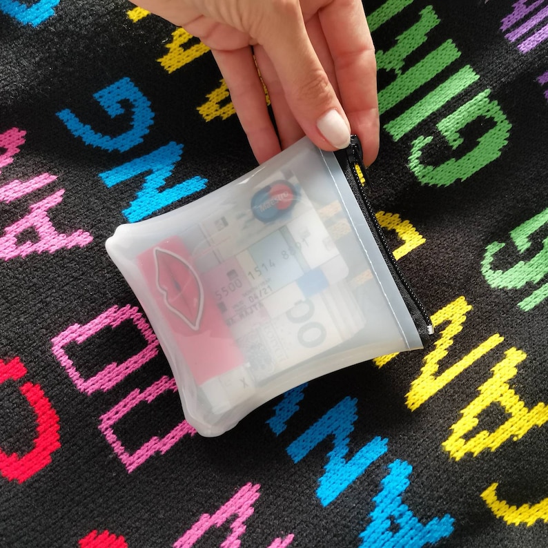 Puede incluir: Una bolsa transparente de pl&aacute;stico con cremallera, con una pegatina de labios rosa y varios billetes de euro en su interior. La bolsa est&aacute; sobre una tela negra con letras de colores.