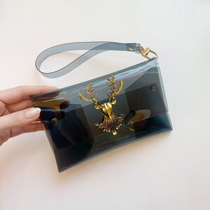 May include: A clear black iridescent clutch bag with a gold deer head accent. The bag has a detachable strap.