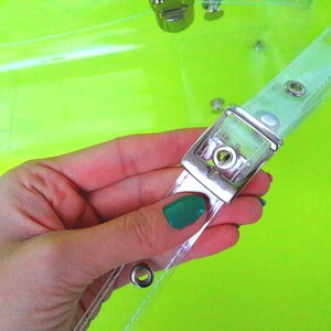 May include: Close-up of a clear plastic bag with a silver buckle and metal eyelets. The bag is held in a hand with a green nail polish.