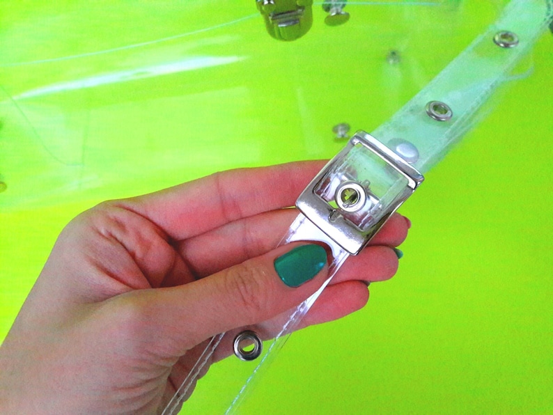 May include: Close-up of a clear plastic bag with a silver buckle and metal eyelets. The bag is held in a hand with a green nail polish.