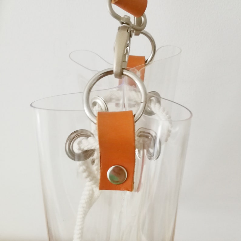 May include: A clear plastic bucket bag featuring tan leather tabs secured with silver metal snaps. The bag includes silver metal rings and a white braided rope drawstring closure, suspended by a silver metal swivel clasp and a tan leather strap.