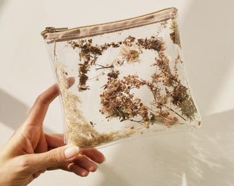 Puede incluir: Una bolsa rectangular transparente llena de flores y hierbas secas. La bolsa tiene una cremallera y un ribete beige. Las plantas secas son de varios tonos de marrón y beige, creando una estética natural.