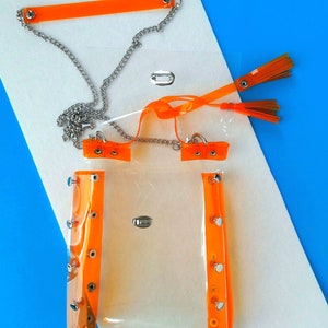 May include: A clear, rectangular handbag with bright orange trim and accents. The bag features a silver chain strap and orange tassels. The trim is secured with silver rivets. The bag is displayed on a white and blue background.