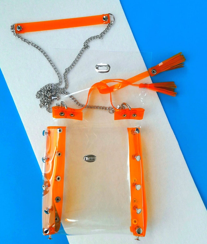 May include: A clear, rectangular handbag with bright orange trim and accents. The bag features a silver chain strap and orange tassels. The trim is secured with silver rivets. The bag is displayed on a white and blue background.