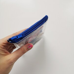 Pode incluir: Uma bolsa transparente e retangular com um z&iacute;per azul vibrante. A bolsa parece conter pequenos itens coloridos. A bolsa &eacute; segurada contra um fundo branco.