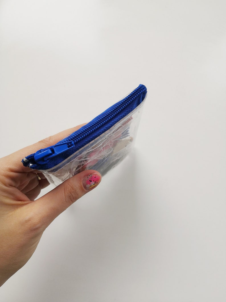 Pode incluir: Uma bolsa transparente e retangular com um z&iacute;per azul vibrante. A bolsa parece conter pequenos itens coloridos. A bolsa &eacute; segurada contra um fundo branco.