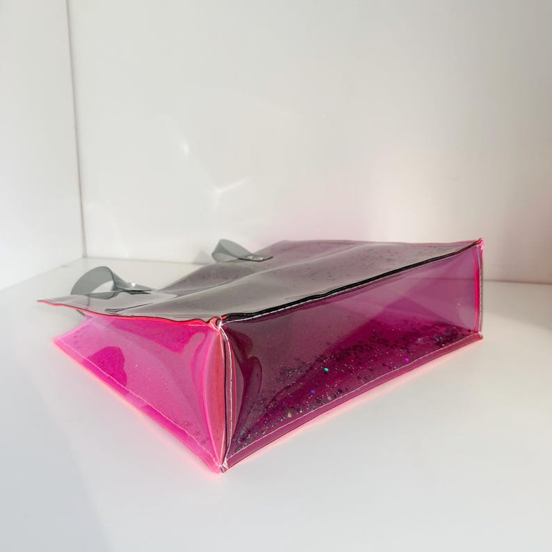 Puede incluir: Un bolso tote transparente con un degradado de tonos rosa y morado. El bolso tiene forma rectangular con una base brillante y asas grises. El bolso est&aacute; hecho de un material transparente y flexible.