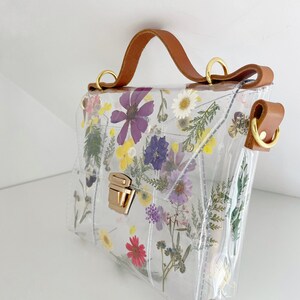 May include: A clear plastic handbag with a brown leather strap and a gold clasp. The bag is decorated with pressed flowers in shades of purple, pink, yellow, and white.