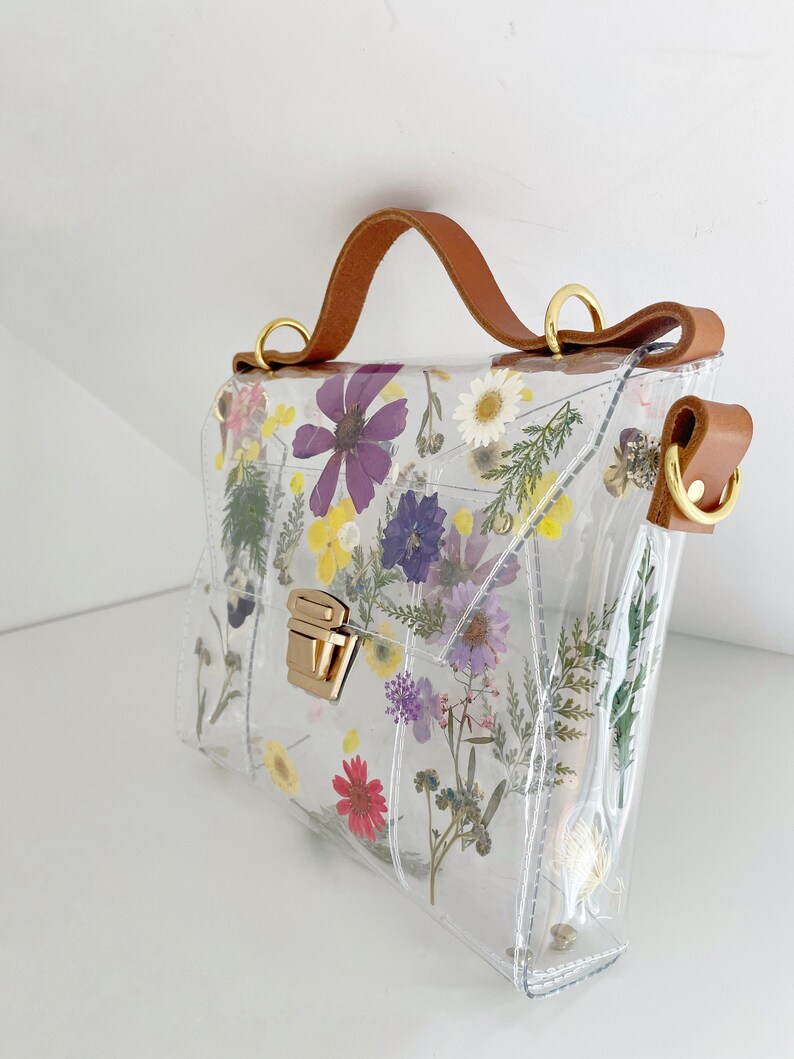 May include: A clear plastic handbag with a brown leather strap and a gold clasp. The bag is decorated with pressed flowers in shades of purple, pink, yellow, and white.