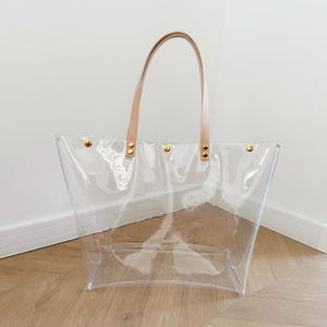 May include: A clear tote bag with tan leather handles and gold-colored rivets. The bag is made of transparent material, allowing visibility of the contents. The handles arch upwards, providing a comfortable grip. The bag is designed for carrying items.