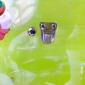 May include: A clear plastic bag with a silver clasp and a silver stud. The bag is on a green background.