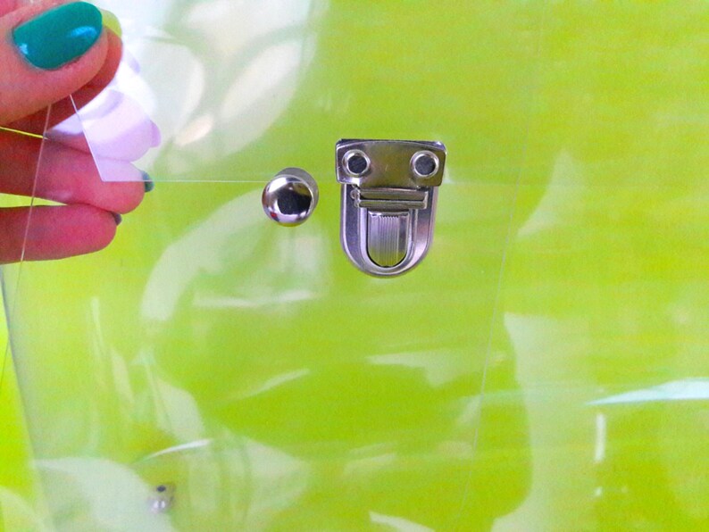 May include: A clear plastic bag with a silver clasp and a silver stud. The bag is on a green background.