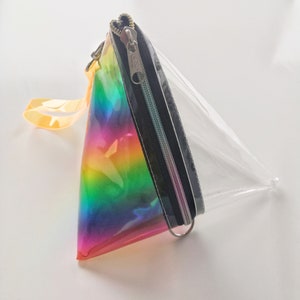 May include: A clear, triangular-shaped pouch with a rainbow iridescent design on one side. The pouch has a black zipper and a silver zipper pull. The pouch has a yellow strap.