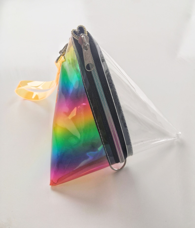 May include: A clear, triangular-shaped pouch with a rainbow iridescent design on one side. The pouch has a black zipper and a silver zipper pull. The pouch has a yellow strap.