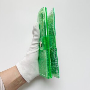 May include: A transparent green wallet with a snap closure. The wallet is filled with glitter and held by a gloved hand. The wallet is made of a clear, shiny material.