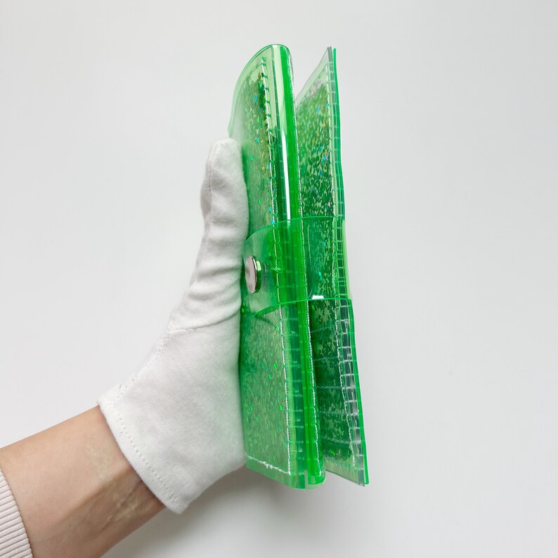 May include: A transparent green wallet with a snap closure. The wallet is filled with glitter and held by a gloved hand. The wallet is made of a clear, shiny material.