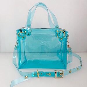 May include: A transparent, turquoise handbag with a rectangular shape. It features a top handle, a detachable shoulder strap, and gold-colored hardware, including rings and a buckle. The bag is made of a clear, flexible material.