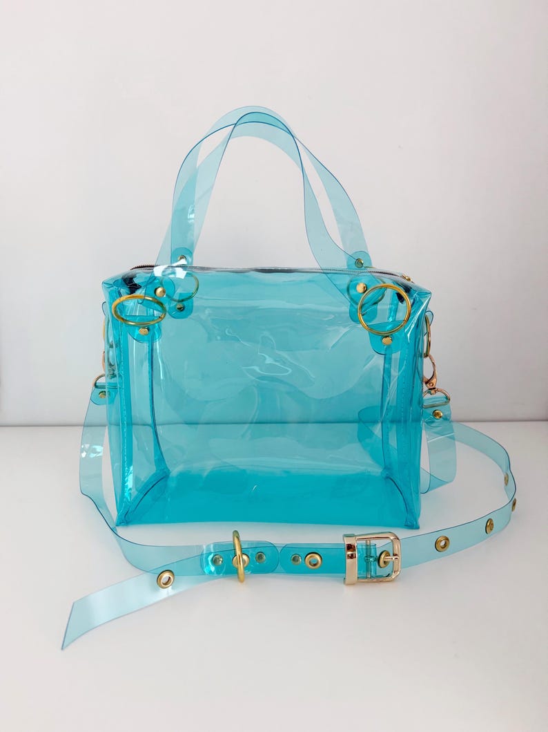May include: A transparent, turquoise handbag with a rectangular shape. It features a top handle, a detachable shoulder strap, and gold-colored hardware, including rings and a buckle. The bag is made of a clear, flexible material.