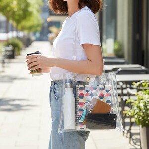 K&ouml;nnte beinhalten: Eine transparente Tragetasche mit einer wei&szlig;en Wasserflasche, einem Notizbuch, einer Brieftasche, einem Telefon und Schl&uuml;sseln. Das Notizbuch hat ein geometrisches Muster in Rot, Blau und Gelb. Die Tasche wird von einer Person getragen, die ein wei&szlig;es T-Shirt und blaue Jeans tr&auml;gt.