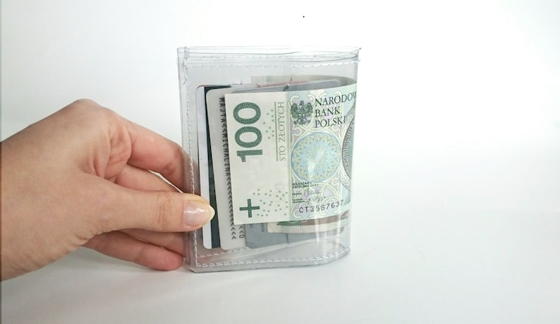 May include: A clear, transparent plastic wallet containing a 100 Polish Zloty banknote and several credit cards. The wallet features visible stitched edges and a rectangular shape, allowing the contents to be clearly seen through the material.