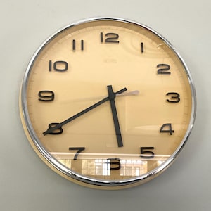 Könnte beinhalten: Eine runde Wanduhr mit cremefarbenem Zifferblatt und schwarzen Ziffern. Die Uhr hat einen silberfarbenen Metallrand und schwarze Zeiger. Die Uhrzeit ist ungefähr 5:38 Uhr. Die Uhr ist an einer hellgrauen Wand befestigt.