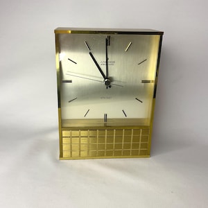 May include: A gold-toned Junghans ATO-MAT clock with a rectangular face and a grid-patterned base. The clock face has black hands and minimalist hour markers. The text "JUNHANS electronic" and "ATO-MAT" are visible on the face. The clock is made in Germany.
