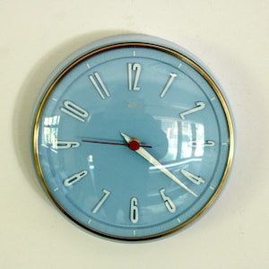Puede incluir: Un reloj de pared redondo con esfera azul claro, números blancos y un borde dorado. El reloj cuenta con manecillas plateadas para las horas y los minutos, un segundero rojo fino y una pequeña tapa central roja bajo un cristal.