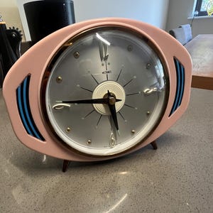May include: An oval-shaped Metamec table clock with a pale pink frame, a silver face, and black hands. The clock features blue accent stripes on the sides, a circular white center detail, and small gold-toned hour markers under a glass cover.