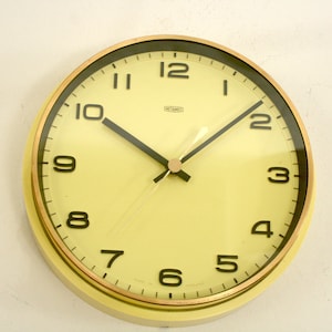 Puede incluir: Un reloj de pared redondo con esfera de color amarillo pálido, números arábigos negros y manecillas negras finas. El reloj presenta un borde metálico en tono dorado y un pequeño logotipo rectangular cerca del centro superior que dice Metamec.