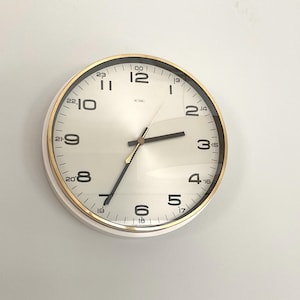 Könnte beinhalten: Eine runde Wanduhr mit einem metallisch goldenen Rahmen und einem weißen Zifferblatt. Das Zifferblatt zeigt schwarze Ziffern für das 12- und 24-Stunden-Format. Sie hat drei dünne Zeiger, darunter einen goldenen Sekundenzeiger, und das Wort METAME ist oben mittig in Schwarz aufgedruckt.