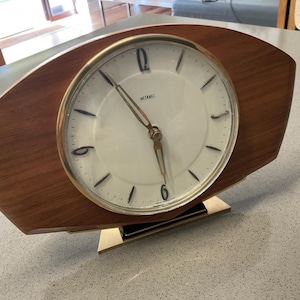 May include: A Metamec mantel clock with a curved wooden frame and a circular white face. The clock features a gold-toned metal bezel, black hour markers, and black hands. It is supported by a rectangular gold-toned metal base with a black accent.