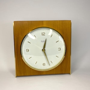 May include: A square wooden clock with a gold-tone bezel and a white clock face. The clock face has black numerals and gold-tone hands. The clock is labeled "Junghans" and "ATO-MAT" on the face.