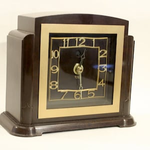 May include: A dark brown mantle clock with a stepped, geometric wooden frame. The square clock face features a gold-toned border, gold-colored Arabic numerals from one to twelve, and two thin, pointed gold-colored hands set against a dark background.