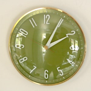 Puede incluir: Un reloj de pared redondo con marco de metal dorado y esfera de color verde oliva brillante. El reloj presenta números arábigos blancos, manecillas blancas para las horas y los minutos, y un segundero rojo fino bajo una lente de cristal.